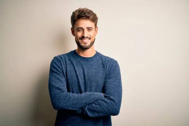 Photo of Young handsome man with beard wearing casual sweater standing over white background happy face smiling with crossed arms looking at the camera. Positive person.