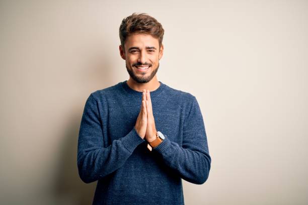 istock Young handsome man with beard wearing casual sweater standing over white background praying with hands together asking for forgiveness smiling confident. 1212702123