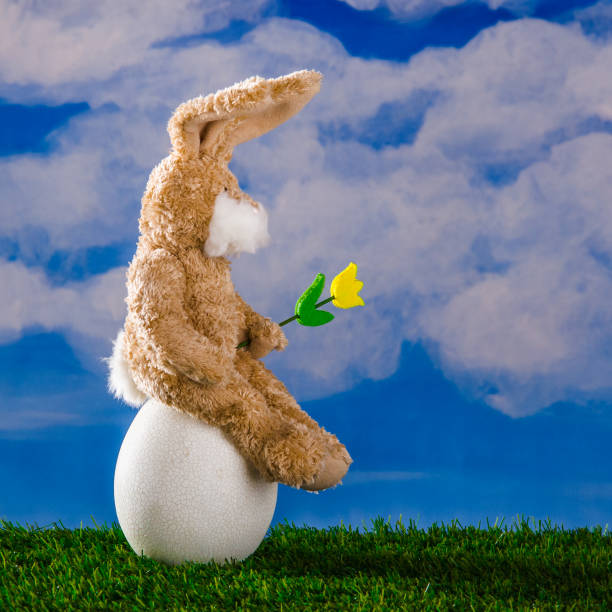 istock Funny rabbit with yellow tulip sitting on a large easter egg. Spring happy sunny day, blue sky,green grass. 1212703362