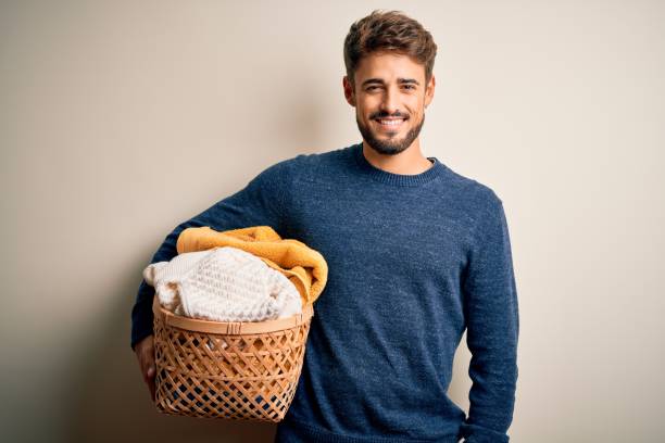 jeune homme faisant le panier d’osier de fixation de ménage avec des vêtements au-dessus du fond blanc avec un visage heureux restant et souriant avec un sourire confiant affichant des dents - toothy smile relaxation white healthcare and medicine photos et images de collection