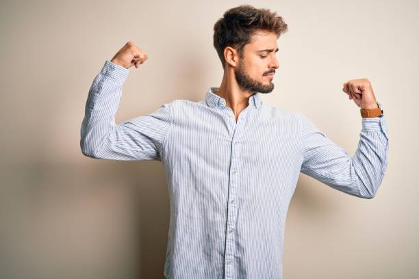 istock Young handsome man with beard wearing striped shirt standing over white background showing arms muscles smiling proud. Fitness concept. 1212703832