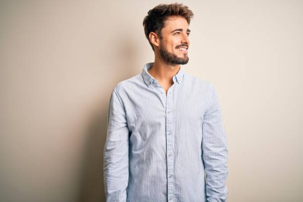 istock Young handsome man with beard wearing striped shirt standing over white background looking away to side with smile on face, natural expression. Laughing confident. 1212703838