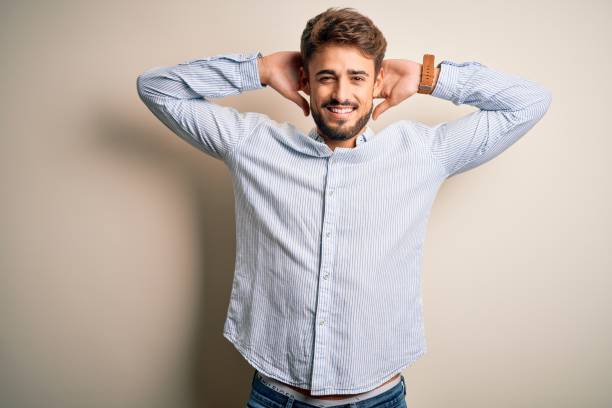 Young handsome man with beard wearing striped shirt standing over white background relaxing and stretching, arms and hands behind head and neck smiling happy