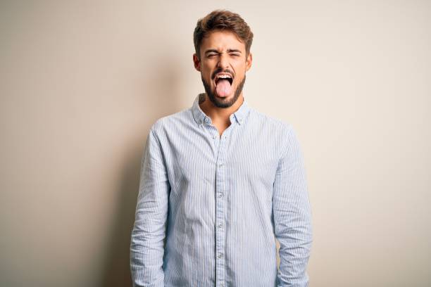 istock Young handsome man with beard wearing striped shirt standing over white background sticking tongue out happy with funny expression. Emotion concept. 1212704469