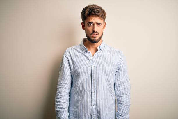 istock Young handsome man with beard wearing striped shirt standing over white background In shock face, looking skeptical and sarcastic, surprised with open mouth 1212704671
