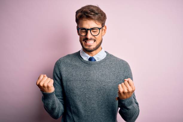 istock Young handsome man with beard wearing glasses and sweater standing over pink background excited for success with arms raised and eyes closed celebrating victory smiling. Winner concept. 1212706005