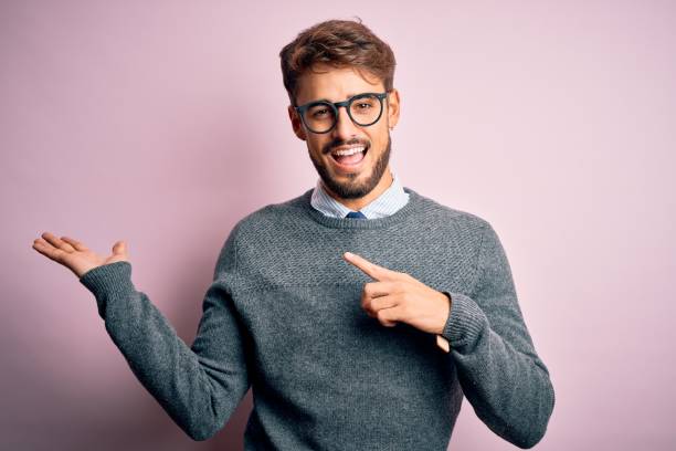 istock Young handsome man with beard wearing glasses and sweater standing over pink background amazed and smiling to the camera while presenting with hand and pointing with finger. 1212706048