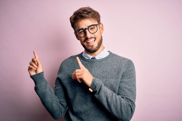 istock Young handsome man with beard wearing glasses and sweater standing over pink background smiling and looking at the camera pointing with two hands and fingers to the side. 1212706077