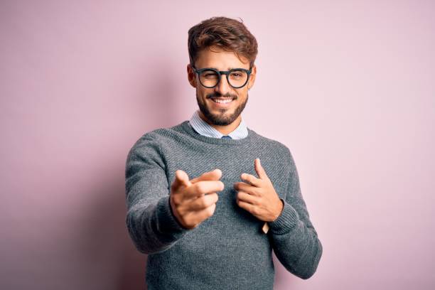 istock Young handsome man with beard wearing glasses and sweater standing over pink background pointing fingers to camera with happy and funny face. Good energy and vibes. 1212706091