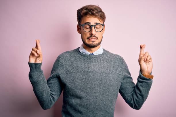 istock Young handsome man with beard wearing glasses and sweater standing over pink background gesturing finger crossed smiling with hope and eyes closed. Luck and superstitious concept. 1212706093