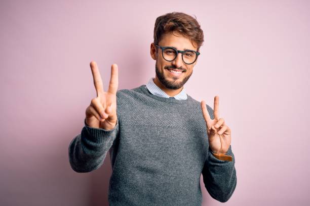 istock Young handsome man with beard wearing glasses and sweater standing over pink background smiling looking to the camera showing fingers doing victory sign. Number two. 1212706099