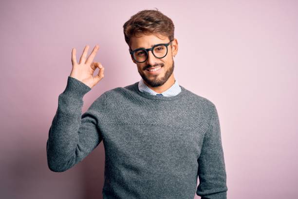 Young handsome man with beard wearing glasses and sweater standing over pink background smiling positive doing ok sign with hand and fingers. Successful expression.