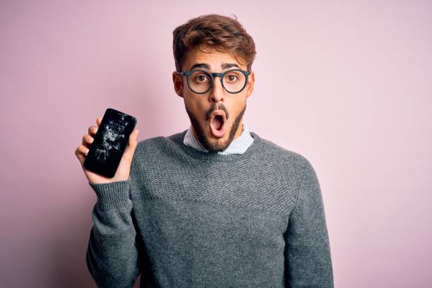 Young man with beard wearing glasses holding broken and craked smartphone scared in shock with a surprise face, afraid and excited with fear expression