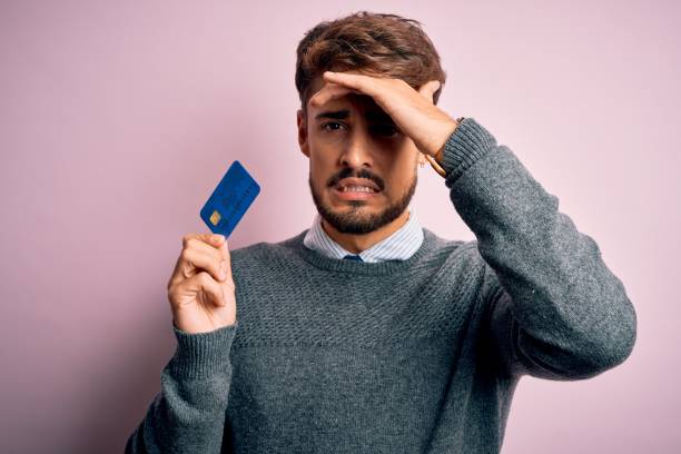 istock Young customer man with beard holding credit card for payment over pink background stressed with hand on head, shocked with shame and surprise face, angry and frustrated. Fear and upset for mistake. 1212706510