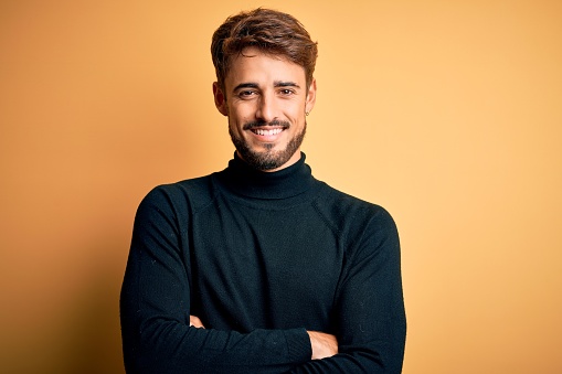 Young handsome man with beard wearing turtleneck sweater standing over yellow background happy face smiling with crossed arms looking at the camera. Positive person.