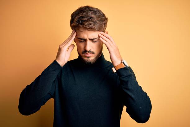 istock Young handsome man with beard wearing turtleneck sweater standing over yellow background with hand on head for pain in head because stress. Suffering migraine. 1212708145