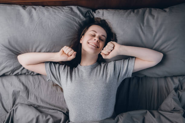 Top view of happy glad woman stretches hands, laughs happily as sees pleasant dreams, keeps eyes shut, dressed in casual clothes. Smiling girl enjoying lazy weekend morning in comfortable bed at home la vista superior de la mujer feliz feliz se estira las manos, se ríe felizmente como ve sueños agradables, mantiene los ojos cerrados, vestido con ropa casual. chica sonriente disfrutando de la mañana de fin de semana perezoso en la cama cómoda en cas