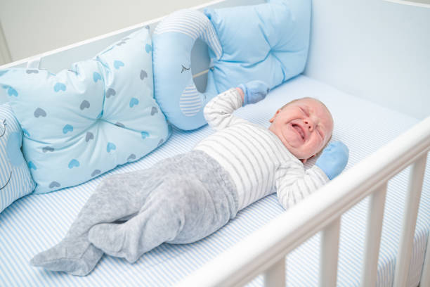 Cute baby lying in his crib and crying. The baby is 1 month old newborn boy of caucasian ethnicity. The scene is situated indoors in family bedroom. The picture is taken with Sony A7III camera.