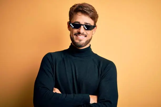 Young man wearing thug life fanny sunglasses standing over isolated yellow background happy face smiling with crossed arms looking at the camera. Positive person.