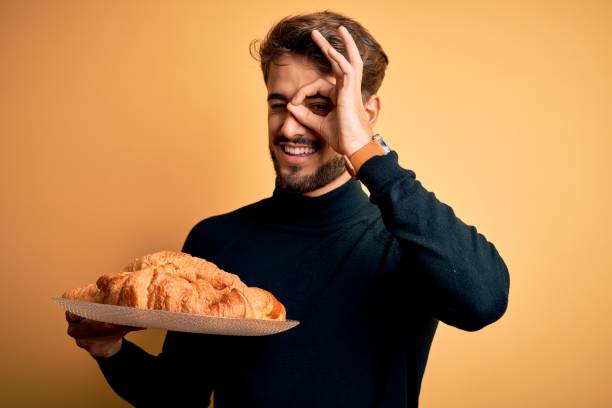 istock Young man with beard holding plate with croissants standing over isolated yellow background with happy face smiling doing ok sign with hand on eye looking through fingers 1212708357