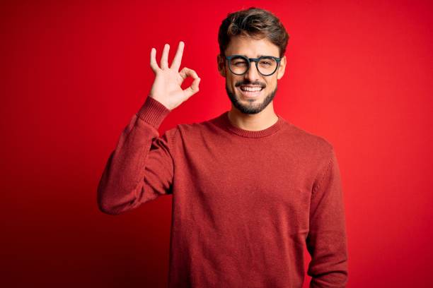 istock Young handsome man with beard wearing glasses and sweater standing over red background smiling positive doing ok sign with hand and fingers. Successful expression. 1212709161