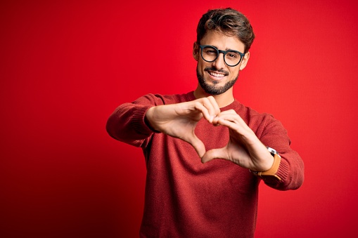 Young handsome man with beard wearing glasses and sweater standing over red background smiling in love doing heart symbol shape with hands. Romantic concept.