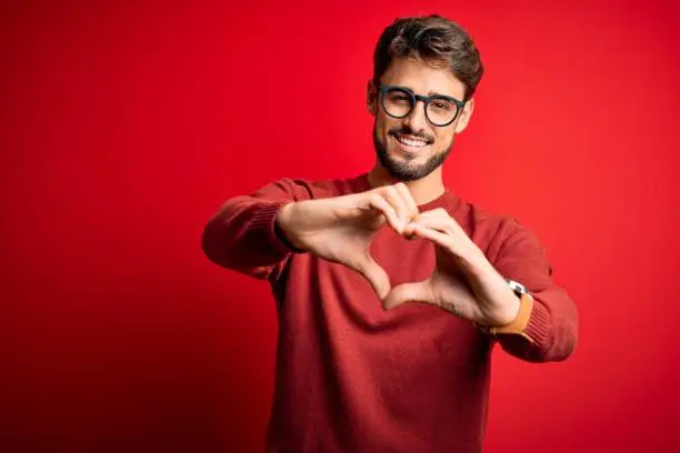 Photo of Young handsome man with beard wearing glasses and sweater standing over red background smiling in love doing heart symbol shape with hands. Romantic concept.