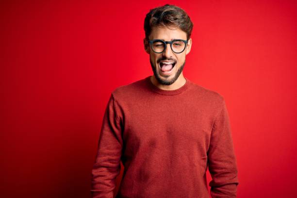 Young handsome man with beard wearing glasses and sweater standing over red background winking looking at the camera with sexy expression, cheerful and happy face. Young handsome man with beard wearing glasses and sweater standing over red background winking looking at the camera with sexy expression, cheerful and happy face. young man wink stock pictures, royalty-free photos & images