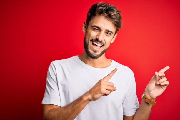 jovem homem bonito com barba vestindo camiseta casual de pé sobre fundo vermelho sorrindo e olhando para a câmera apontando com duas mãos e dedos para o lado. - man pointing - fotografias e filmes do acervo