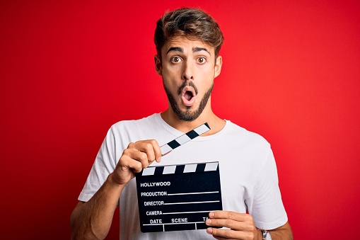 Young director man with beard making movie using clapboard over isolated red background scared in shock with a surprise face, afraid and excited with fear expression