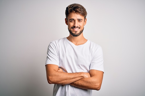 Young handsome man with beard wearing casual t-shirt standing over white background happy face smiling with crossed arms looking at the camera. Positive person.