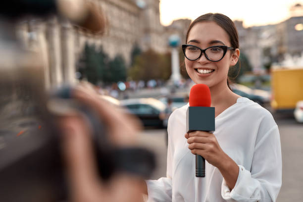 Watch us work for you. TV reporter presenting the news outdoors. Journalism industry, live streaming concept. Portrait of professional female reporter at work. Young woman standing on the street with a microphone in hand and smiling at camera. Camera man recording it. Horizontal shot. Selective focus on woman blouse photos stock pictures, royalty-free photos & images