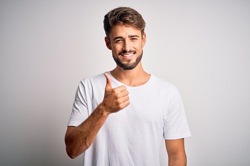 Young handsome man with beard wearing casual t-shirt standing over white background doing happy thumbs up gesture with hand. Approving expression looking at the camera showing success.