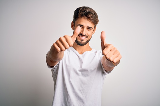 Young handsome man with beard wearing casual t-shirt standing over white background approving doing positive gesture with hand, thumbs up smiling and happy for success. Winner gesture.