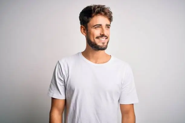 Photo of Young handsome man with beard wearing casual t-shirt standing over white background looking away to side with smile on face, natural expression. Laughing confident.