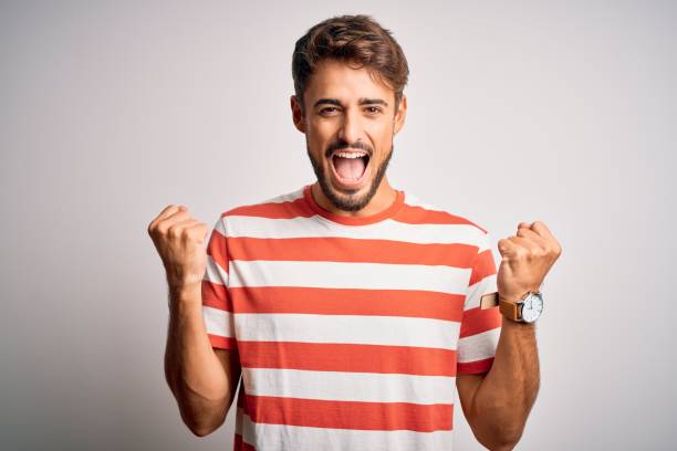 istock Young handsome man with beard wearing striped t-shirt standing over white background celebrating surprised and amazed for success with arms raised and open eyes. Winner concept. 1212711408