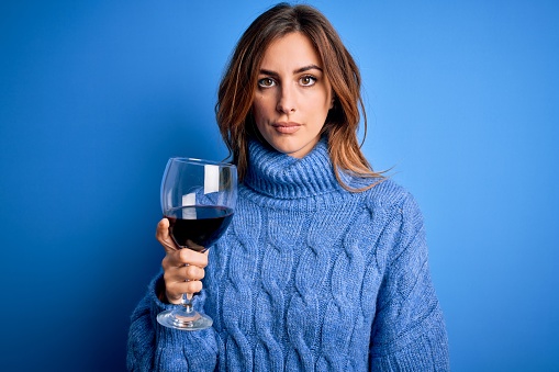 Young beautiful brunette woman drinking glass of red wine over isolated blue background with a confident expression on smart face thinking serious