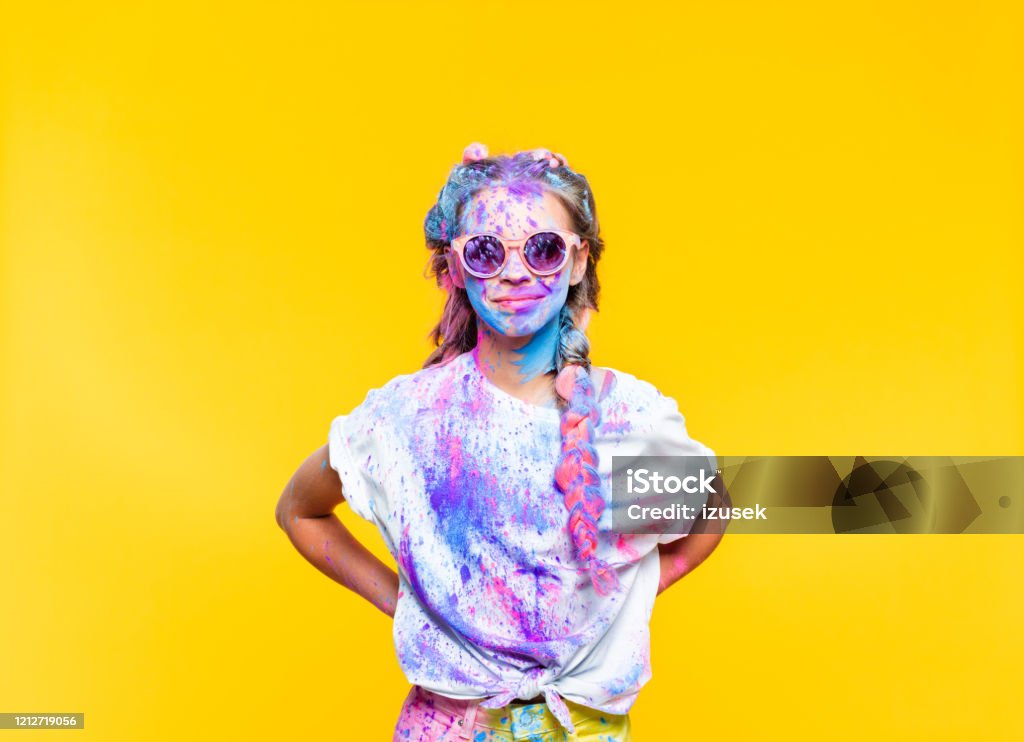 Cheerful teenage girl covered in holi powder Happy teenage girl wearing sunglasses covered in colorful powder after holy festival, smiling at camera. Studio shot, yellow background. Colors Stock Photo