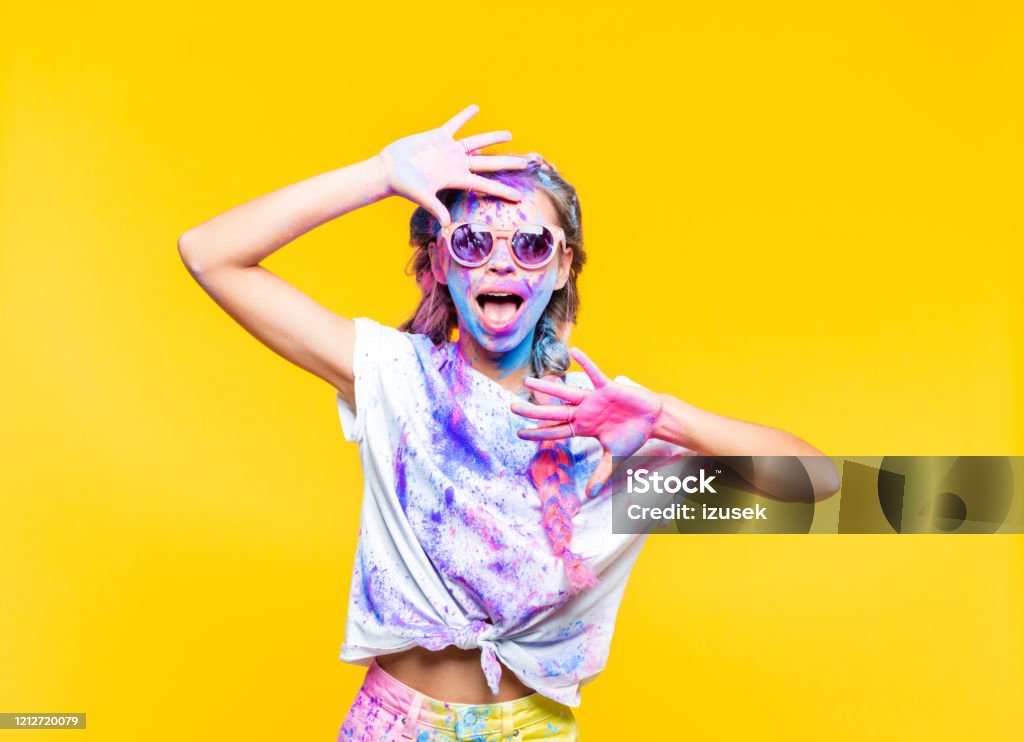Happy holi festival Excited teenage girl wearing sunglasses, covered in colorful powder holy festival, yelling at camera. Studio shot, yellow background. Carefree Stock Photo