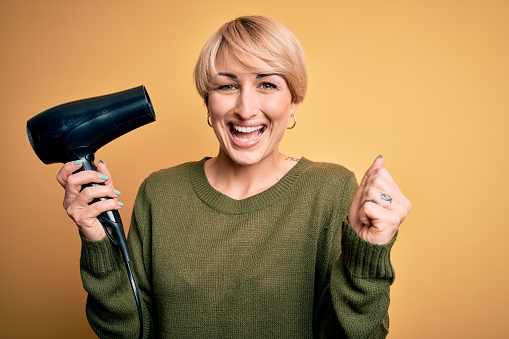 Young blonde woman with short hair drying her hair using hairdryer over yellow background screaming proud and celebrating victory and success very excited, cheering emotion