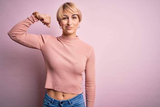 Young blonde woman with short hair wearing casual turtleneck sweater over pink background Strong person showing arm muscle, confident and proud of power Young blonde woman with short hair wearing casual turtleneck sweater over pink background Strong person showing arm muscle, confident and proud of power bicep photos stock pictures, royalty-free photos & images