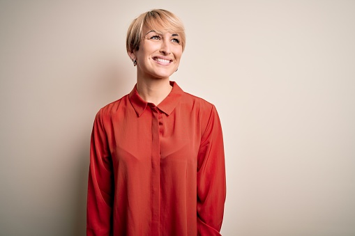 Young beautiful business blonde woman with short hair standing over isolated background looking away to side with smile on face, natural expression. Laughing confident.
