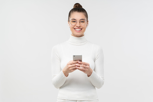 Studio shot of beautiful girl, looking straight at camera, smiling while holding smartphone with both hands, isolated on gray background