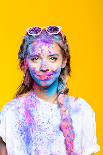Happy teenage girl covered in colorful powder after holy festival, smiling at camera. Studio shot, yellow background.
