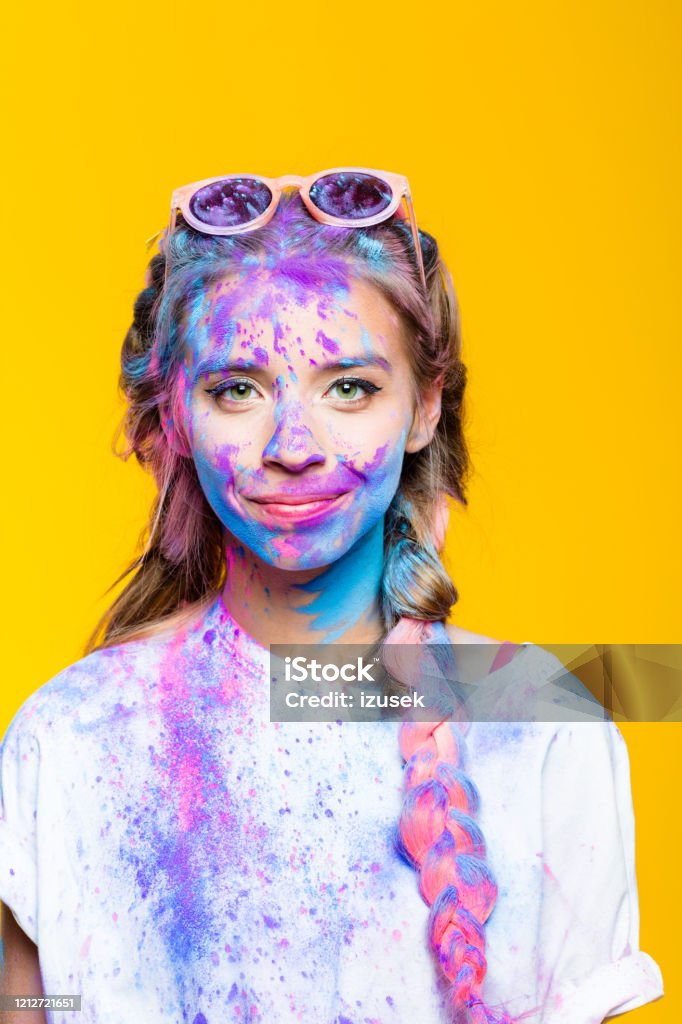 Portrait of cheerful teenage girl celebrating holi festival Happy teenage girl covered in colorful powder after holy festival, smiling at camera. Studio shot, yellow background. Yellow Background Stock Photo