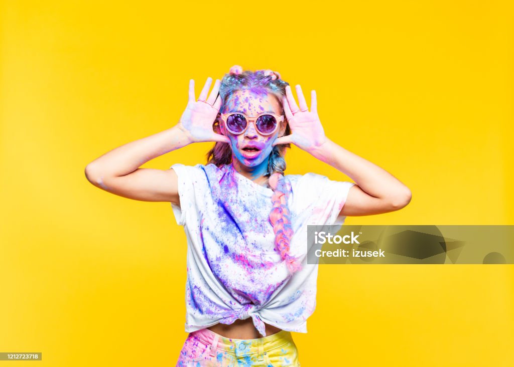 Shocked teenage girl celebrating holi festival Portrait of surprised teenage girl covered in colorful powder after holy festival, yelling at camera. Studio shot, yellow background. Humor Stock Photo