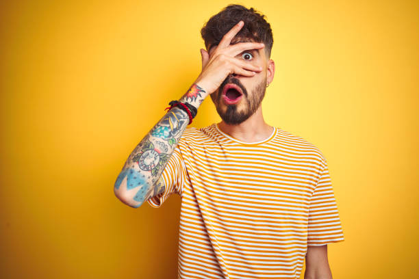 Young man with tattoo wearing striped t-shirt standing over isolated yellow background peeking in shock covering face and eyes with hand, looking through fingers with embarrassed expression. Young man with tattoo wearing striped t-shirt standing over isolated yellow background peeking in shock covering face and eyes with hand, looking through fingers with embarrassed expression. Hidden Meaning stock pictures, royalty-free photos & images