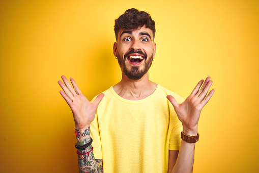 Young man with tattoo wearing t-shirt standing over isolated yellow background celebrating crazy and amazed for success with arms raised and open eyes screaming excited. Winner concept