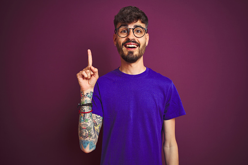 Young man with tattoo wearing t-shirt and glasses standing over isolated purple background pointing finger up with successful idea. Exited and happy. Number one.