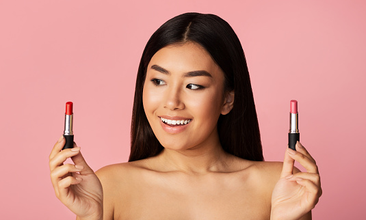 Makeup Concept. Beautiful Asian Girl Holding Two Lipsticks Choosing Lipgloss Standing On Pink Background. Studio Shot